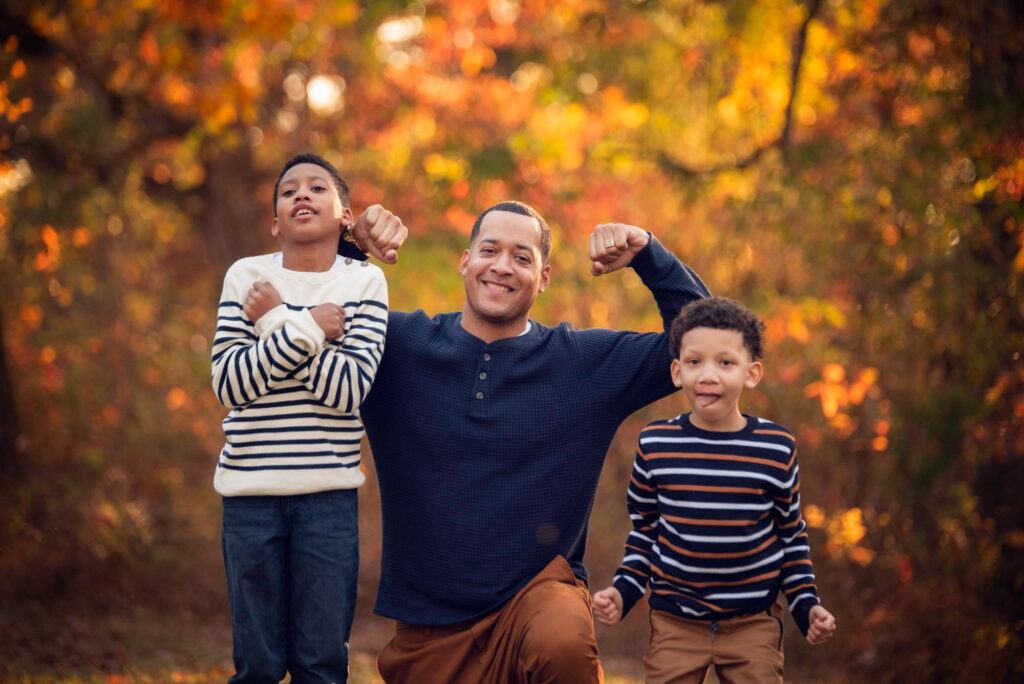 Father flexing with two sons among autumn leaves during a playful fall photo in Birmingham, AL.