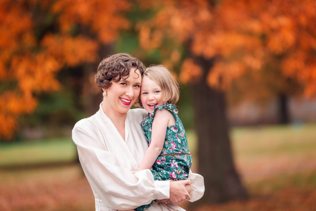 Mom holding her daughter in front of vibrant orange foliage during Birmingham fall photos.