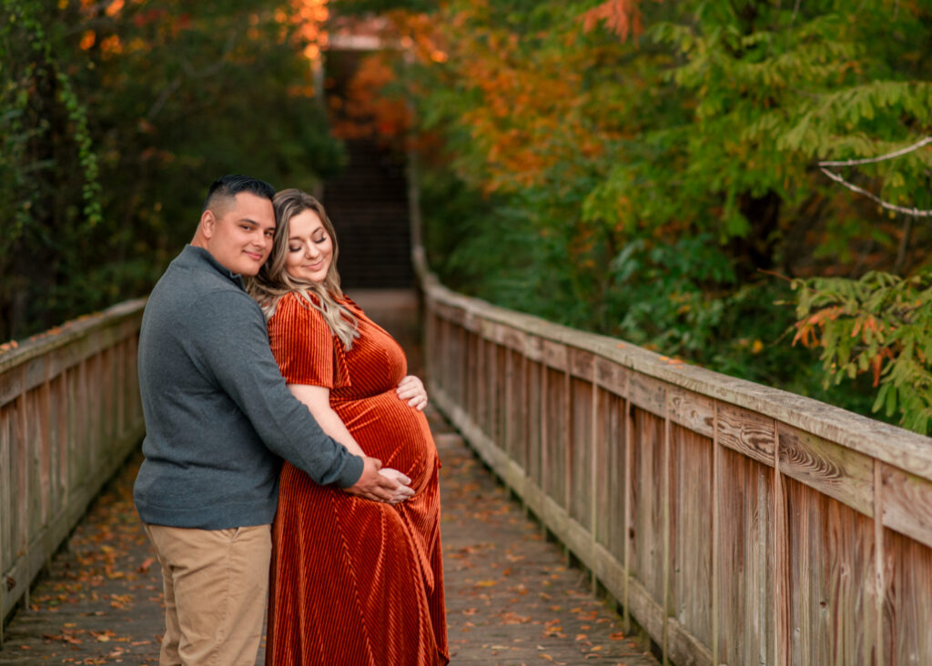 Couple holding each other on bridge with autumn leaves in Birmingham fall maternity mini session.