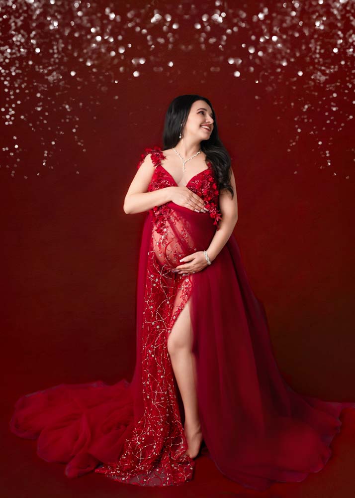 Studio maternity photography in Birmingham, AL—expectant mother in a red sequin gown on a rich burgundy backdrop, hands on belly, smiling.