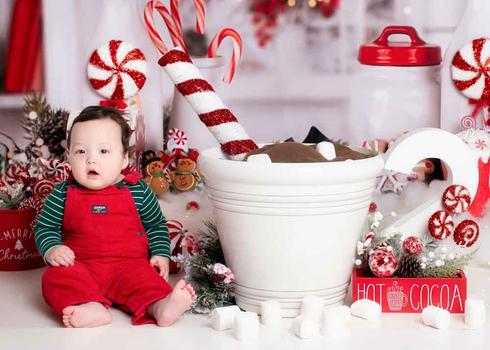 Baby in red overalls beside an oversized hot cocoa mug with marshmallows on a festive peppermint studio set in Birmingham, AL.
