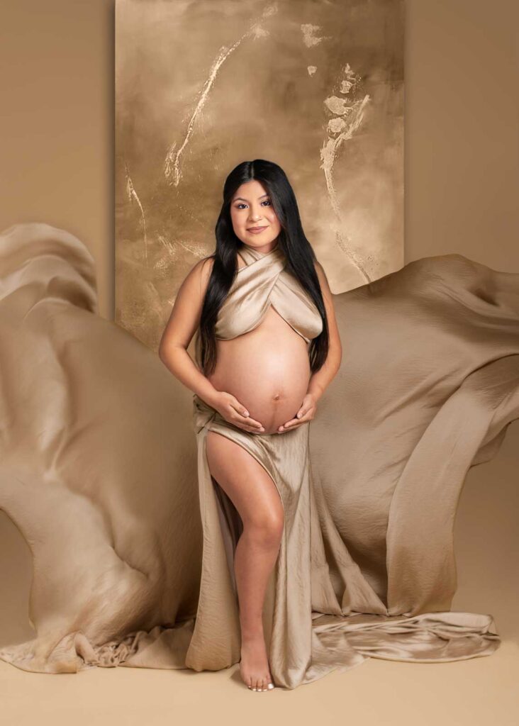 Pregnant mother in flowing taupe gown standing in Birmingham, AL studio maternity photography session with golden backdrop.