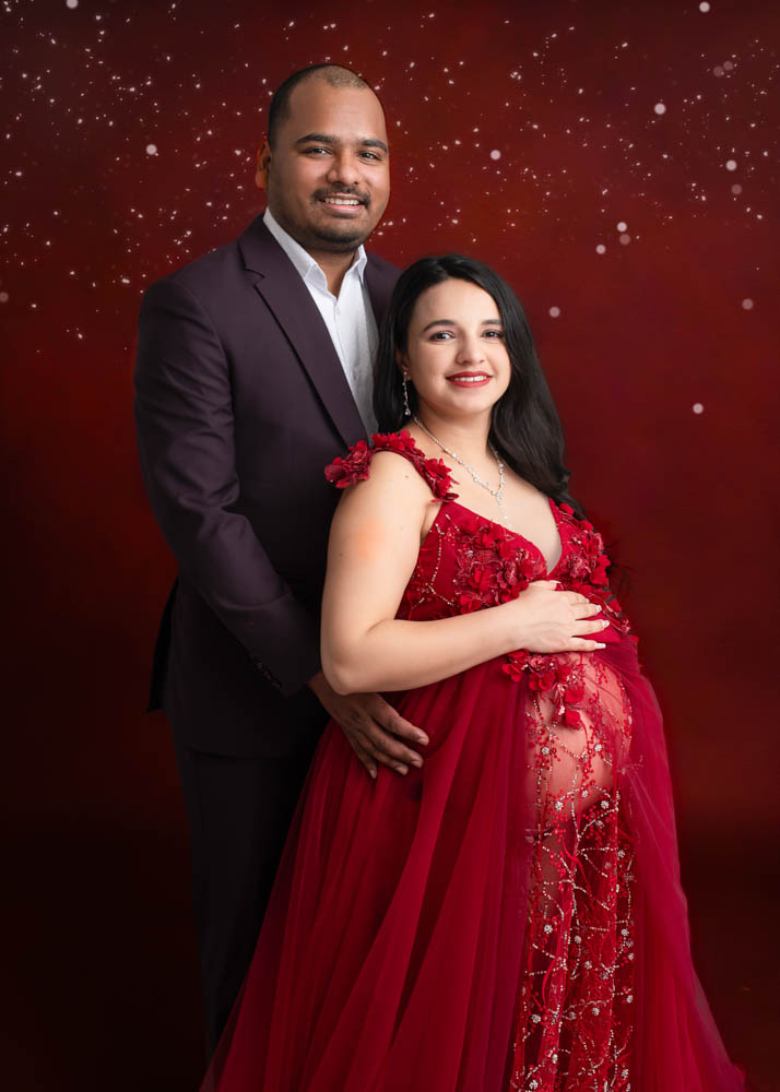 Maternity couple smiling during a studio maternity photography session in Birmingham, featuring a red sequin gown and burgundy backdrop with subtle sparkle effects