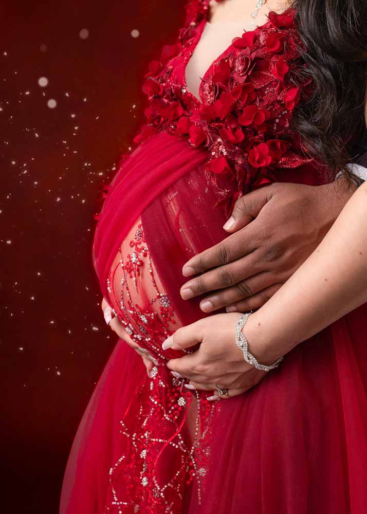 Close-up of expectant parents’ hands on a baby bump, showcasing the red embellished maternity gown during a studio maternity photography session in Birmingham.