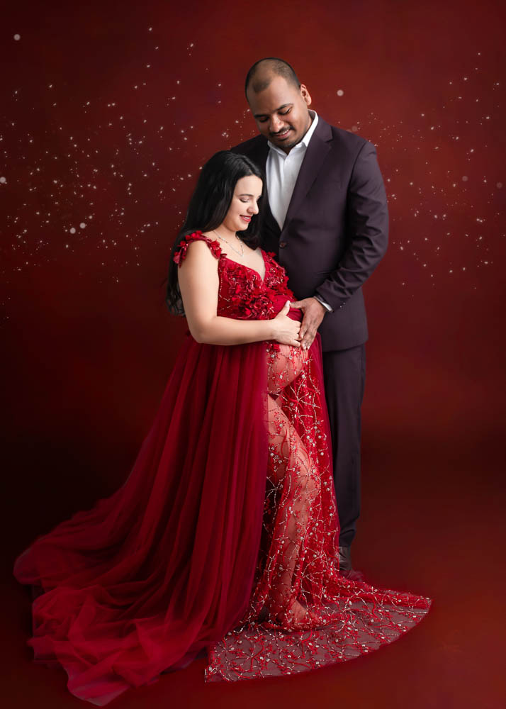 Expectant mother in a red embellished maternity gown standing with her partner in a Birmingham studio, posing against a rich burgundy backdrop with soft sparkling accents.