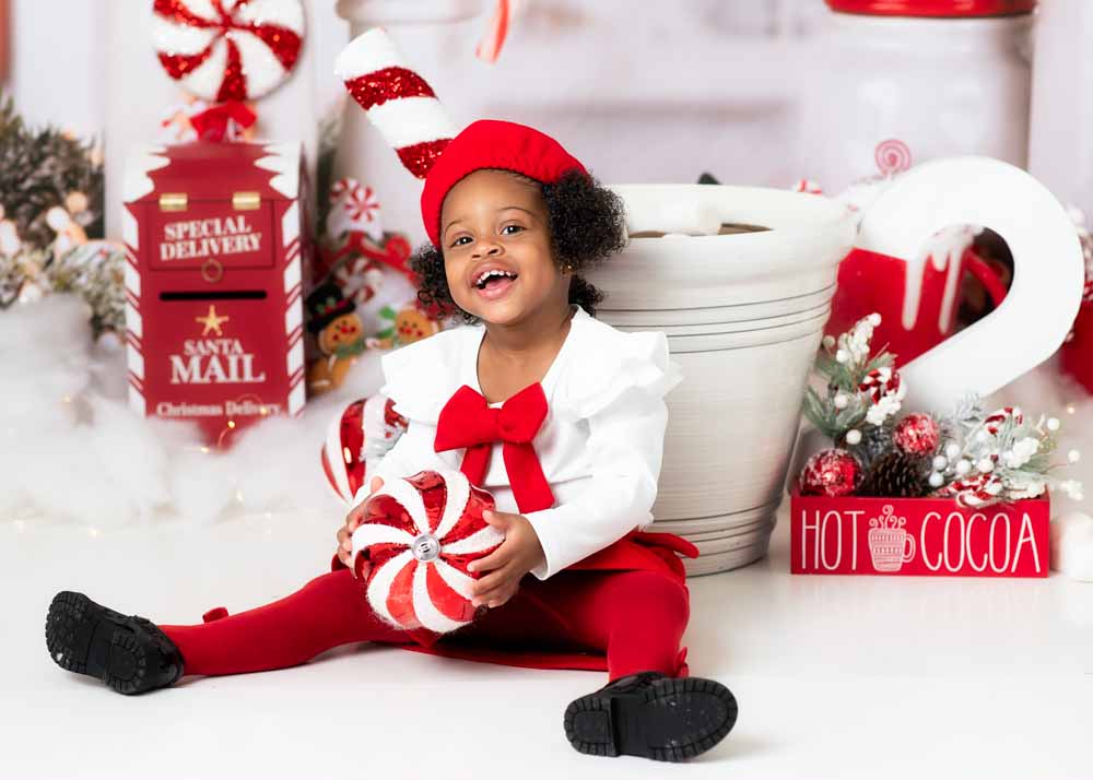 Toddler giggling while seated, holding a large peppermint ornament near the “Hot Cocoa” sign on a studio set, Birmingham, AL.
