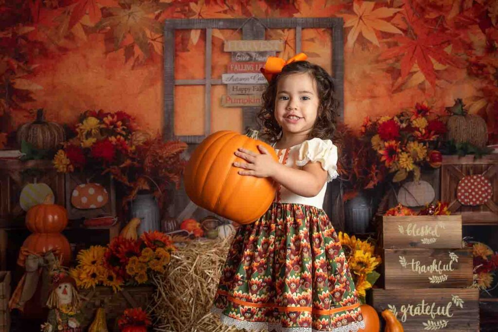 Toddler girl holding pumpkin in festive fall studio milestone set with autumn leaves and seasonal décor in Birmingham, AL photography studio.