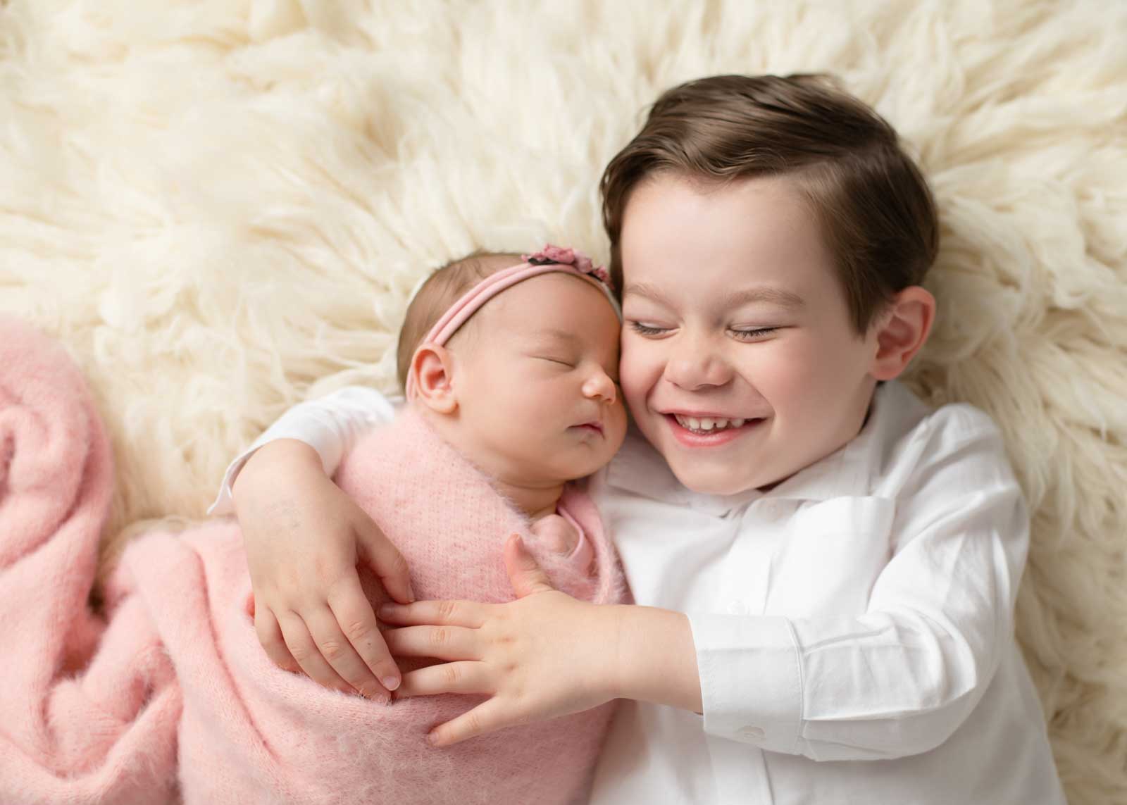 Sibling newborn photos: three-year-old big brother giggles while holding newborn sister Emma on ivory flokati.