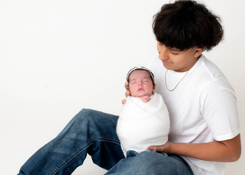 Sibling newborn photos: teen big brother cradles swaddled newborn sister on white backdrop in Birmingham studio.