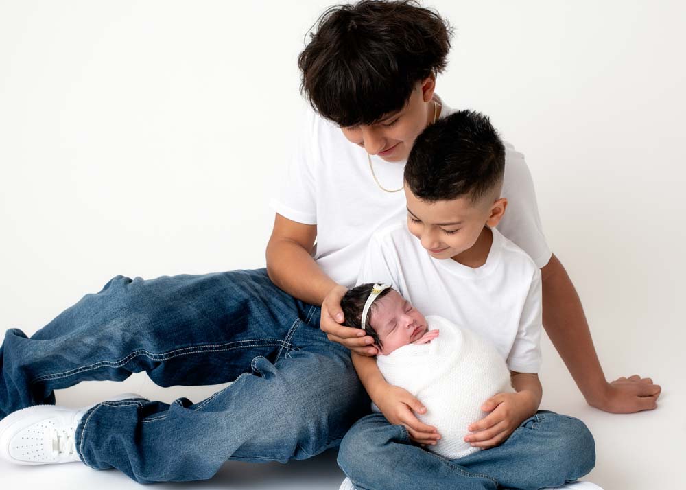 Sibling newborn photos: two big brothers admire their newborn sister, simple white studio portrait.