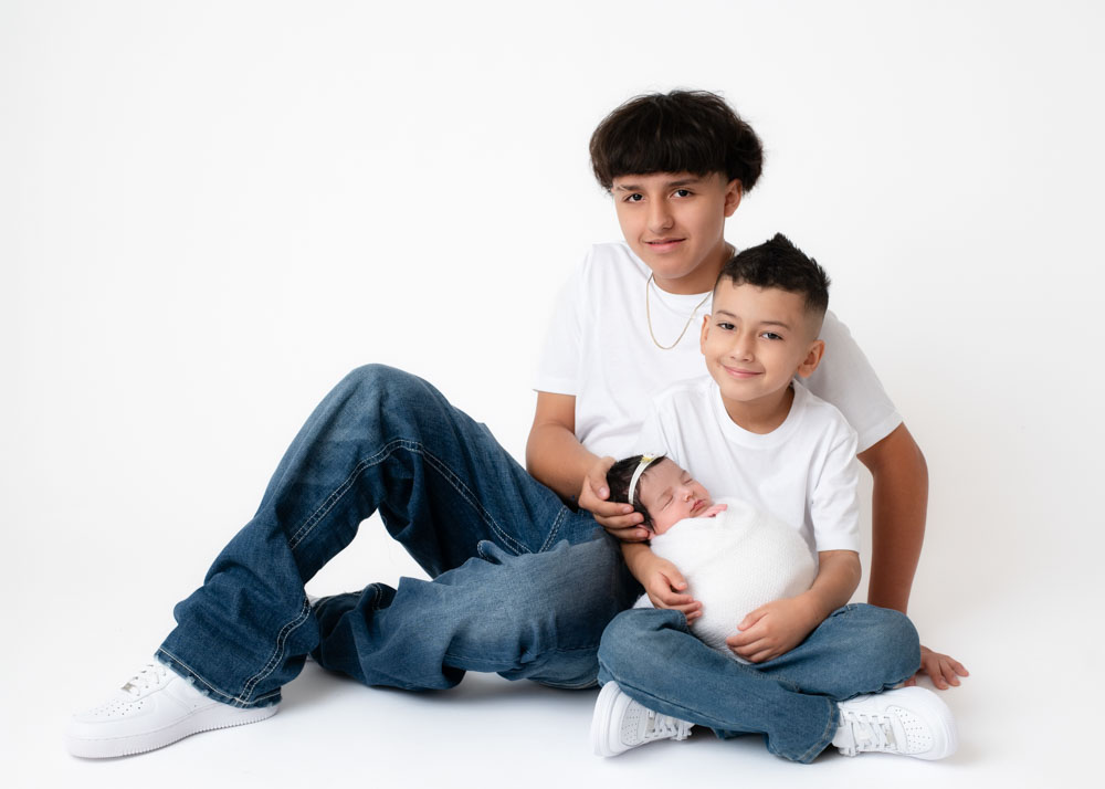 Sibling newborn photos on white: two big brothers seated, holding their newborn sister for a relaxed family portrait.