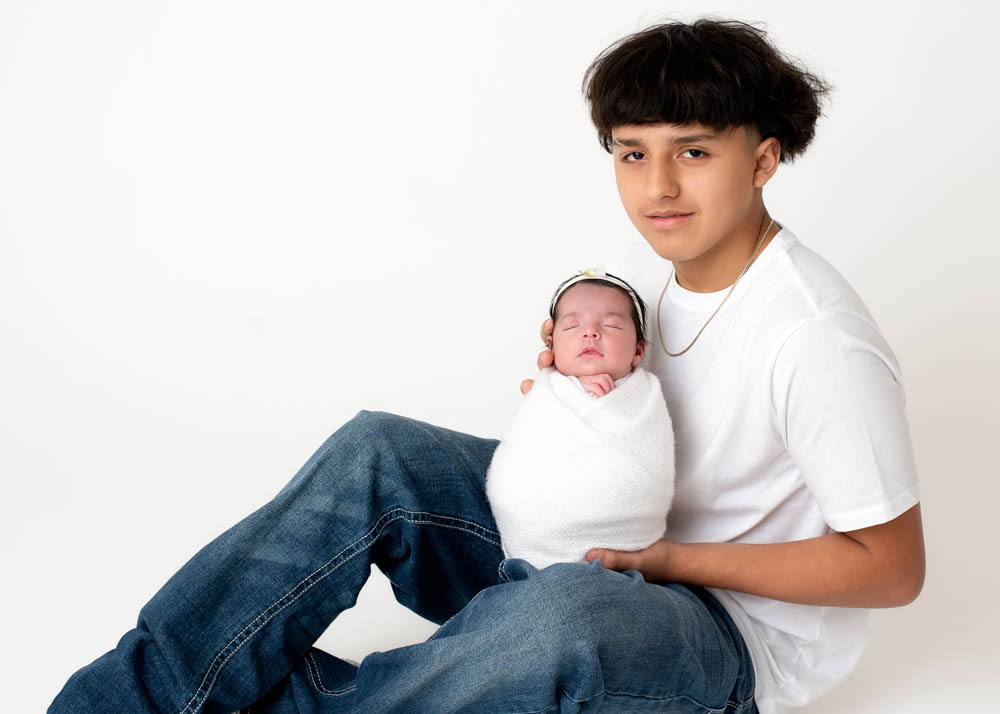 Sibling newborn photos with teen big brother holding newborn sister, clean white studio look.