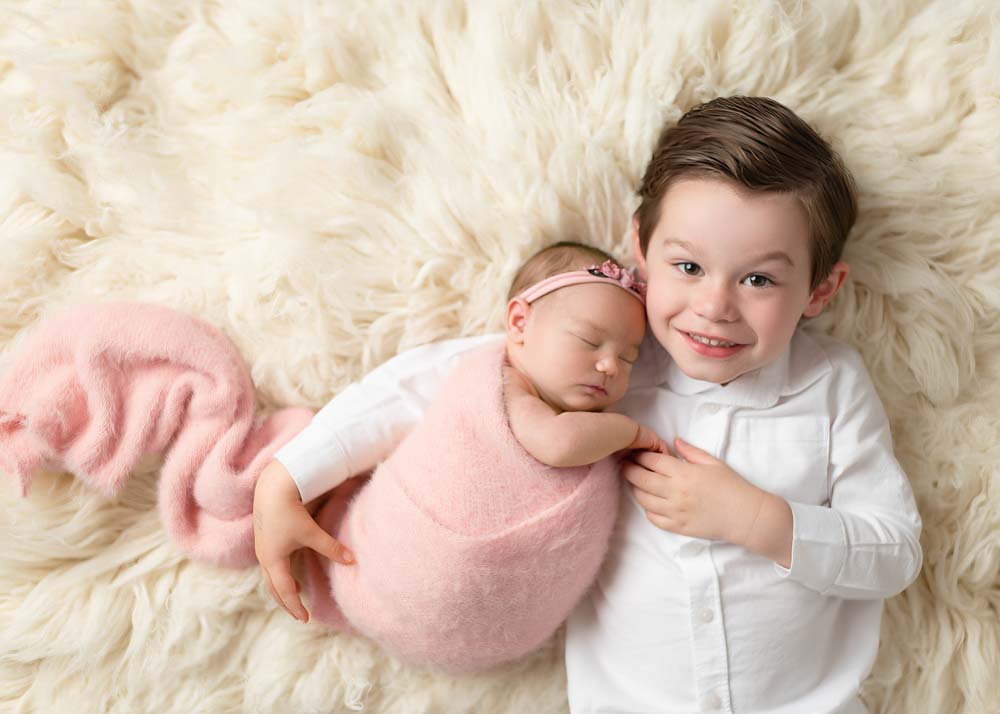 Sibling newborn photos on ivory flokati—big brother reclining with newborn sister for a relaxed portrait.
