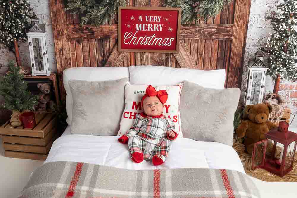 Baby in plaid Christmas pajamas sitting on a rustic holiday bed set with greenery, lanterns, and cozy winter décor in a Birmingham photography studio.