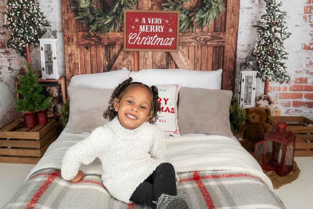 Baby in plaid Christmas pajamas sitting on a rustic holiday bed set with greenery, lanterns, and cozy winter décor in a Birmingham photography studio.