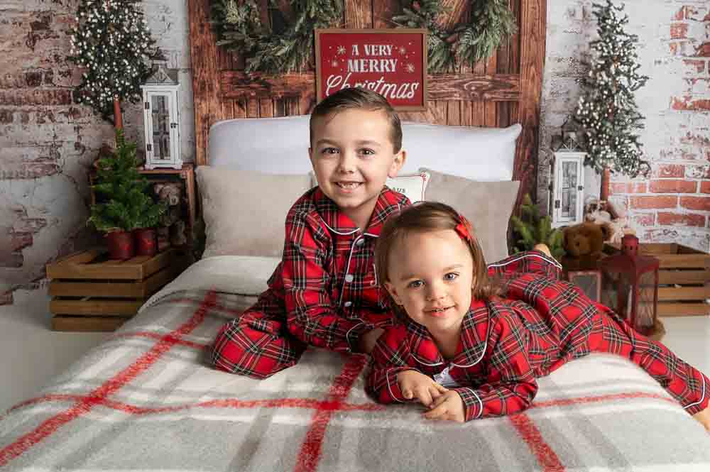 Baby in plaid Christmas pajamas sitting on a rustic holiday bed set with greenery, lanterns, and cozy winter décor in a Birmingham photography studio.