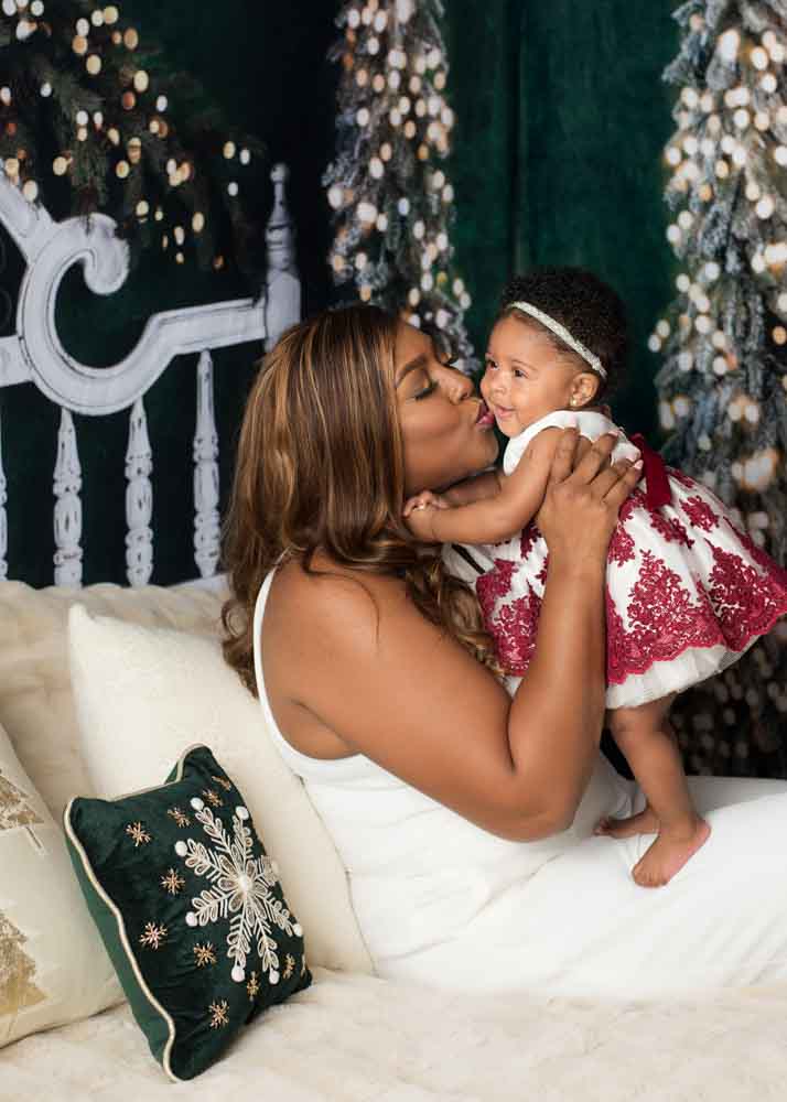 Mother lifting and kissing her baby on the cozy cream Christmas bed with green trees and festive lights in a Birmingham studio.