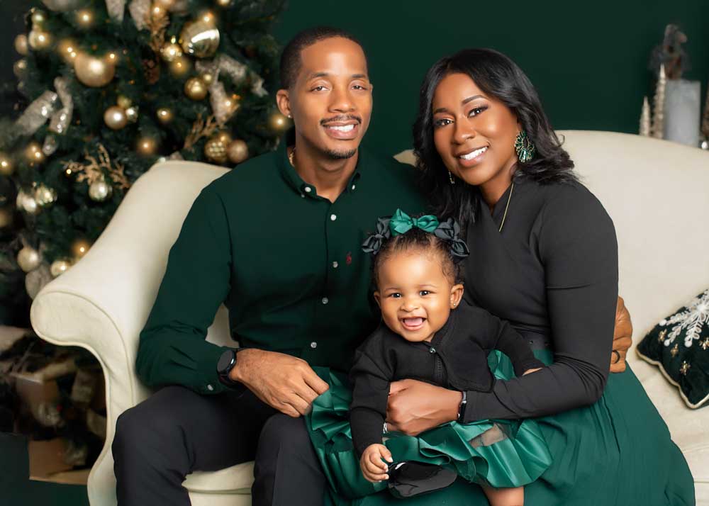 Parents sitting with their toddler on a cream sofa in front of a green and gold Christmas tree during a studio holiday session.