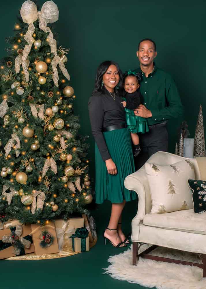 Family of three in coordinated green and black outfits standing beside a gold-decorated Christmas tree in a Birmingham studio.
