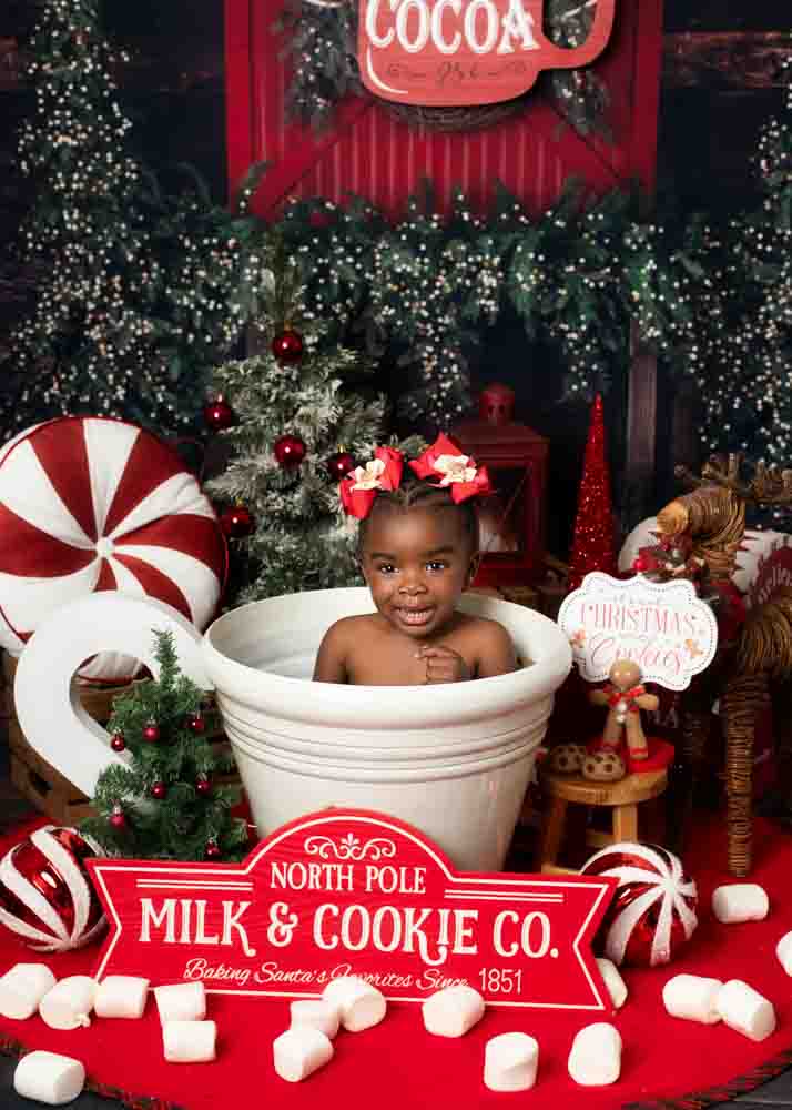 Toddler smiling inside a giant milk-and-cookie mug with marshmallows and peppermint decorations on a Christmas studio set in Birmingham, AL.
