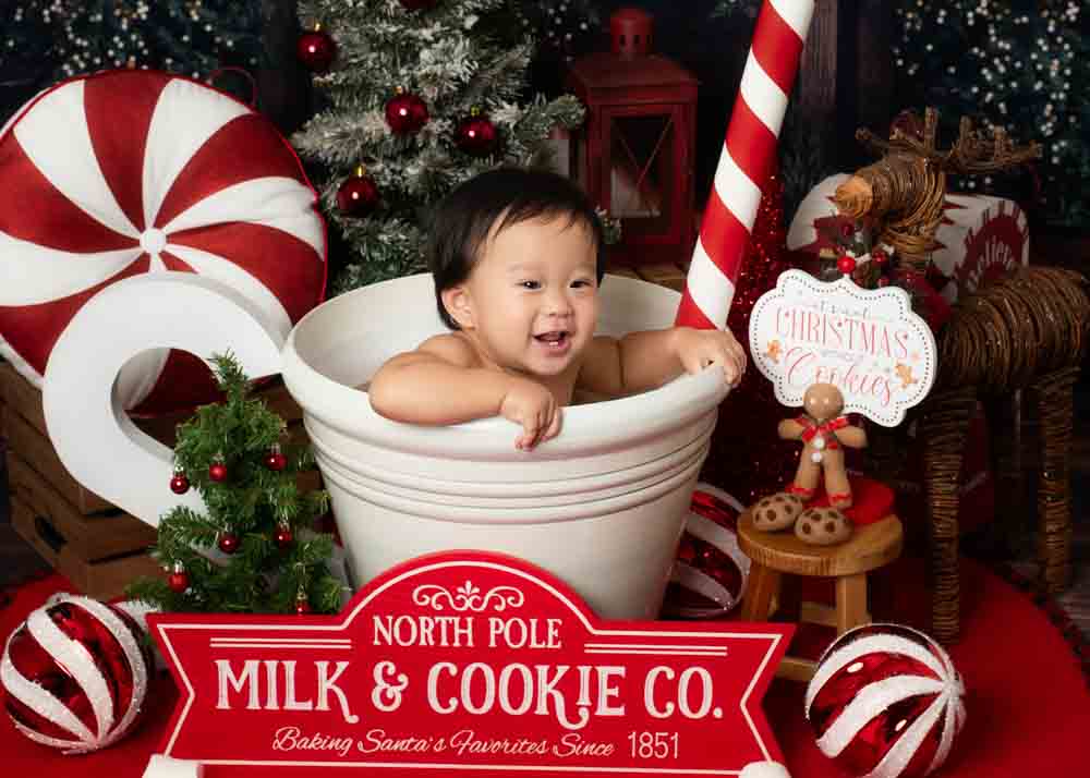 Baby leaning over the edge of a oversized milk-and-cookie mug surrounded by peppermint props and Christmas trees in a Birmingham holiday studio.