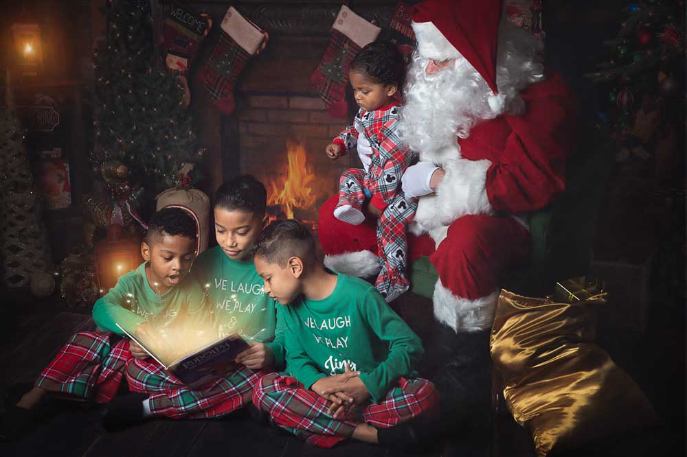 Children sitting by Santa as they read a glowing Christmas storybook in a cozy living room set with fireplace and holiday décor in a Birmingham photography studio