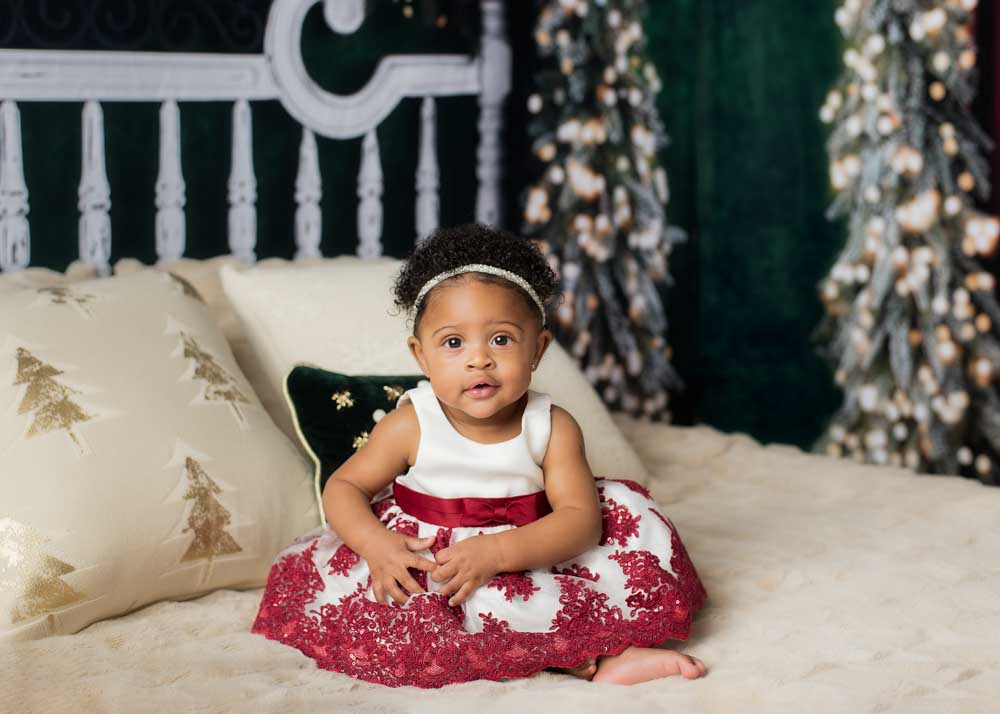 Baby girl sitting on a cozy cream bed with holiday pillows and lit Christmas trees in the background inside a Birmingham photography studio.
