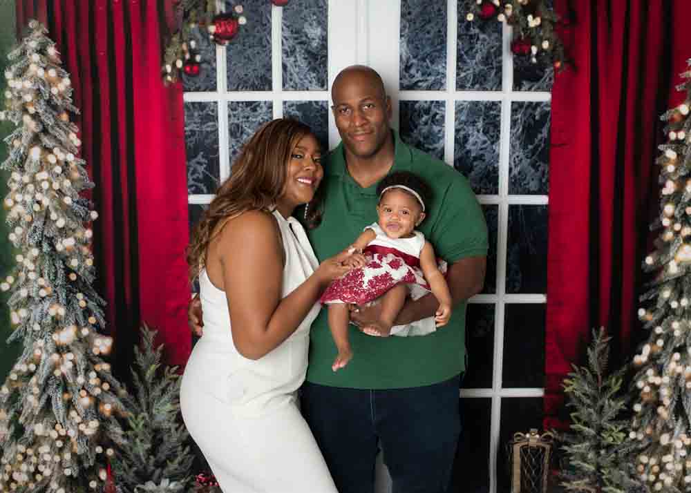 Family standing in front of the snowy window backdrop with red curtains and decorated Christmas trees during a studio holiday session in Birmingham, Alabama.