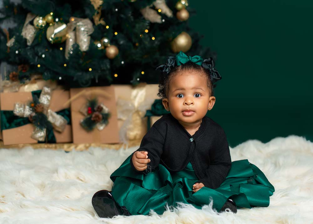 Baby girl in a green holiday dress sitting on a white rug in front of gold-wrapped Christmas presents and a decorated tree in a Birmingham photography studio