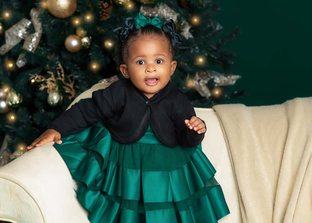 Close-up portrait of a toddler in a green dress smiling in front of a gold Christmas tree.