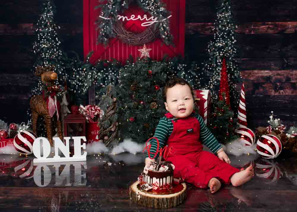 Baby boy in red overalls beside a Christmas cake with peppermint props and evergreen trees.